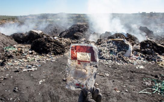 A hand holding up Tesco cheese plastic packaging with burning rubbish heaps in the background.