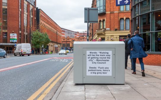 Photo of grey box on Portland Street with notice: "Words: "Walking will be the main way of getting round the city" - Manchester City Council. Deeds: "Fuck you pedestrians, here's a big grey box."