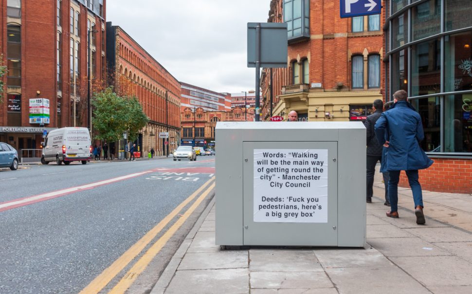 Photo of grey box on Portland Street with notice: "Words: "Walking will be the main way of getting round the city" - Manchester City Council. Deeds: "Fuck you pedestrians, here's a big grey box."