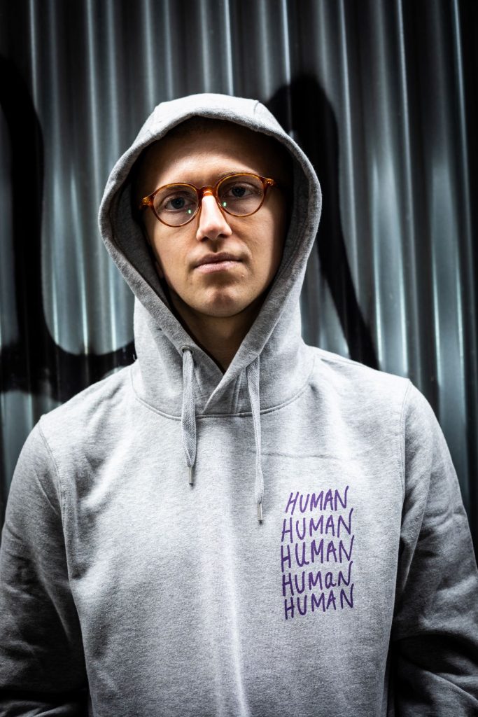 A man wearing glasses and a grey hoodie with Human written 5 times on the upper left.