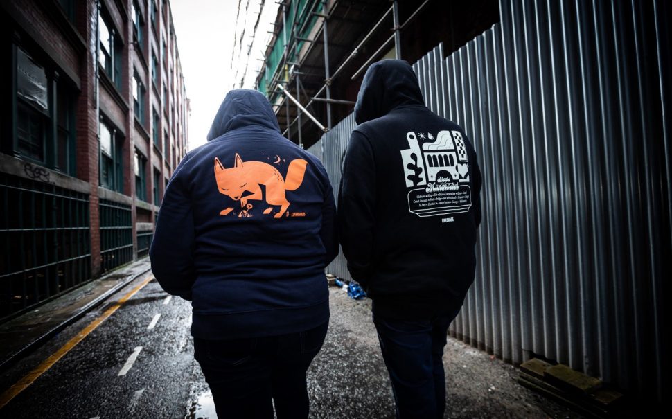 Two people in black hoodies, one with an orange fox, another with a design of Manchester railways.