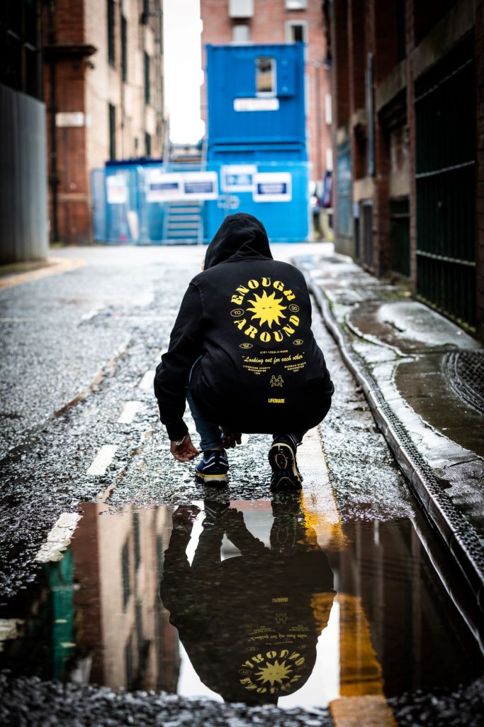 A man crouching on a street wearing a black hoodie with 'Enough To Go Around' written around a sun