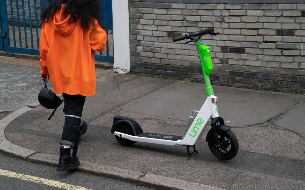 Photo of a woman wearing an orange jacket carrying a helmet. She steps on to the pavement from the road, where there is a Lime e-scooter parked.