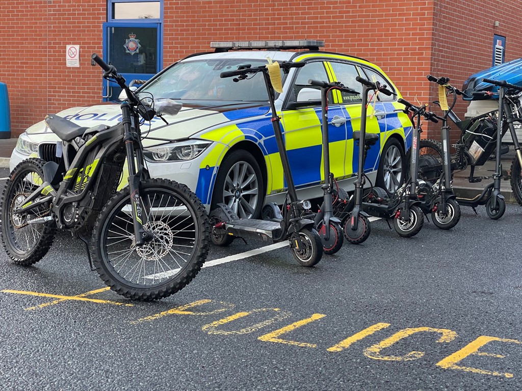 Photo of six black e-scooters in between two Surron electric motorcyles. Yellow tags have been placed on the handlebars of some of the e-scooters. All of them are in front of a police car parked in front of a red brick building with a blue door with a Greater Manchester Police emblem on the window. Yellow markings on the tarmac surface say "POLICE" in capital letters.