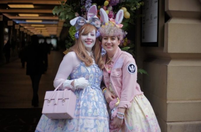 Two people standing in front of an archway wearing sweet lolita fashion. The person on the left is wearing a purple dress and bunny ears and carrying a pink bag. The person on the right is wearing a yellow dress, pink jacket and bunny ears.