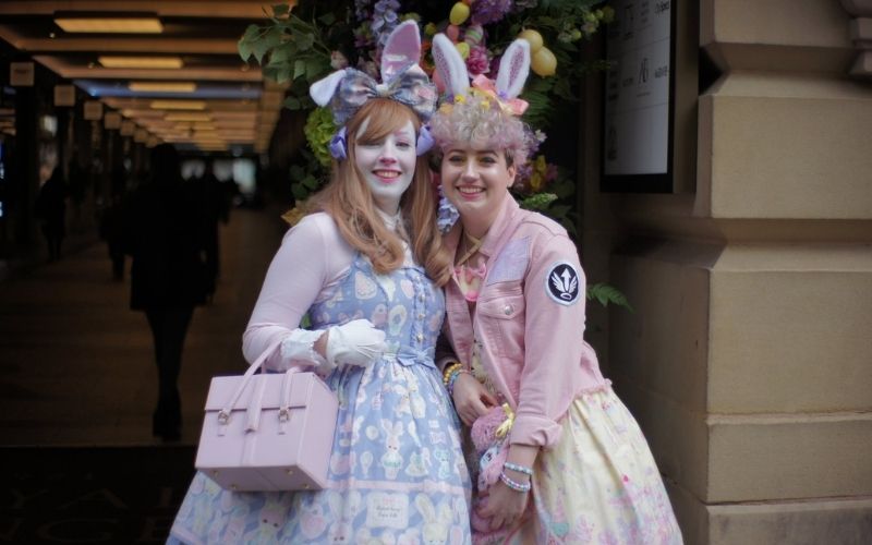 Two people standing in front of an archway wearing sweet lolita fashion. The person on the left is wearing a purple dress and bunny ears and carrying a pink bag. The person on the right is wearing a yellow dress, pink jacket and bunny ears.