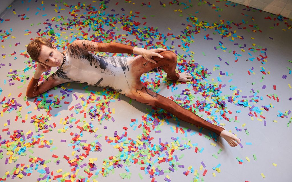 Photo of Linus Karp dressed in a jellicle costume, lying face up on a floor covered covered in confetti.