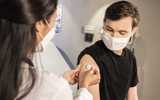 A man, wearing a white cloth face mask has a plaster put on his upper right arm by a woman wearing a white medical respirator mask.