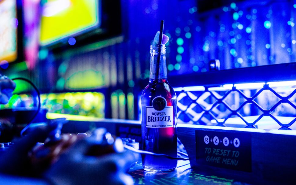 A photograph of a glass bottle with a red liquid inside sat on a bar that is lit with intense blue lighting. The bottle is labelled 'Bowser Breezer'.
