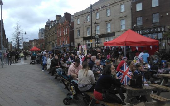 A crowd - sitting and standing - holding and wearing union jacks, watch the coronation