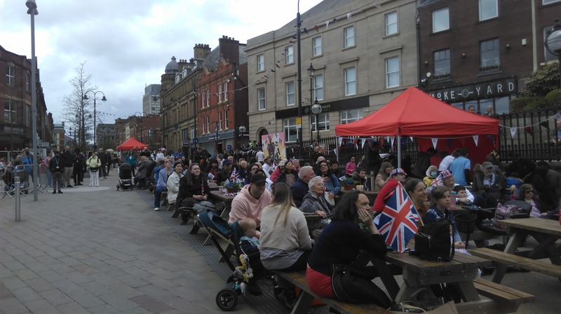 A crowd - sitting and standing - holding and wearing union jacks, watch the coronation