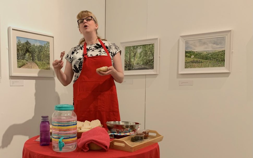 A woman in a red apron performing poetry while spreading hummus on bread, in front of a table of food