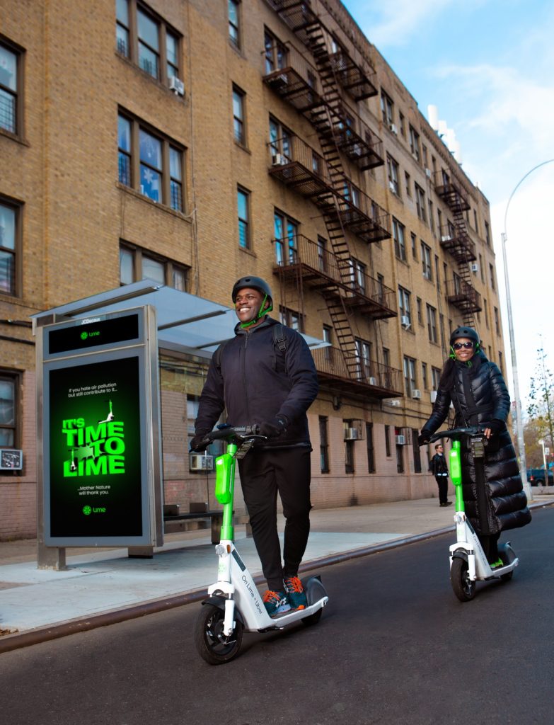 Two people riding on Lime e-scooters next to a bus stop and a block of flats