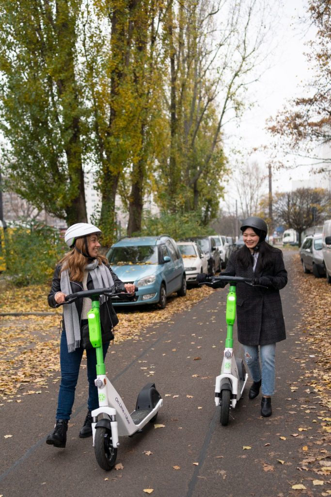 Two people walking with Lime e-bikes on a road with trees and cars in the background