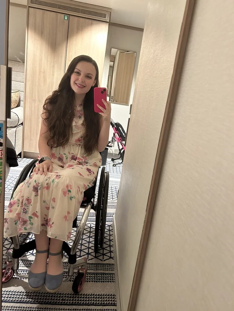 Imali Chislett, 24, taking a selfie with her mobile phone in a mirror. She is sitting in a wheelchair and has long dark hair. She is smiling at the camera. 